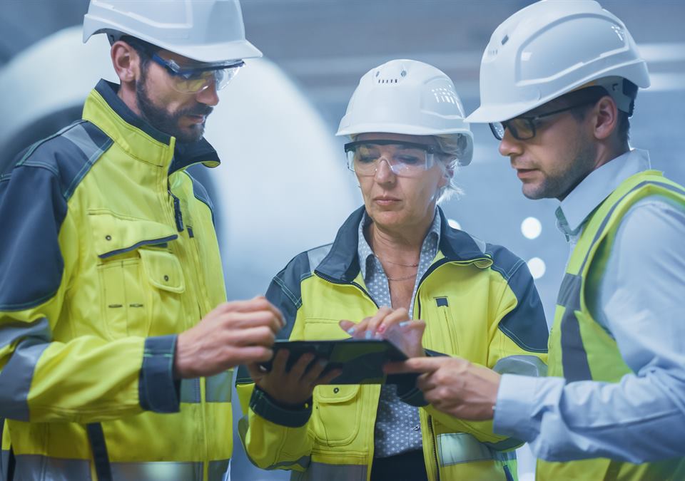 Illustrative stock photo: three engineers in hard hats, safety glasses and high-visibility jackets reviewing work on a tablet